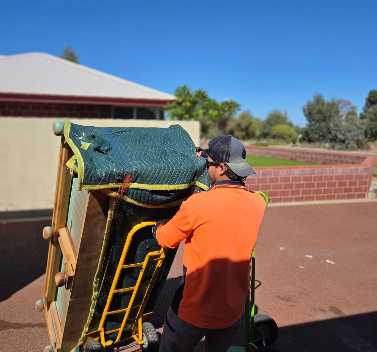 Removalist wheeling wrapped furniture on a trolley past a brick home
